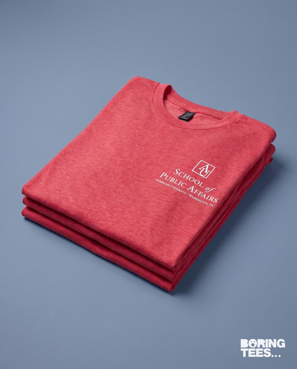 Folded stack of heather red tees with a small white American University School of Public Affairs crest on the left chest