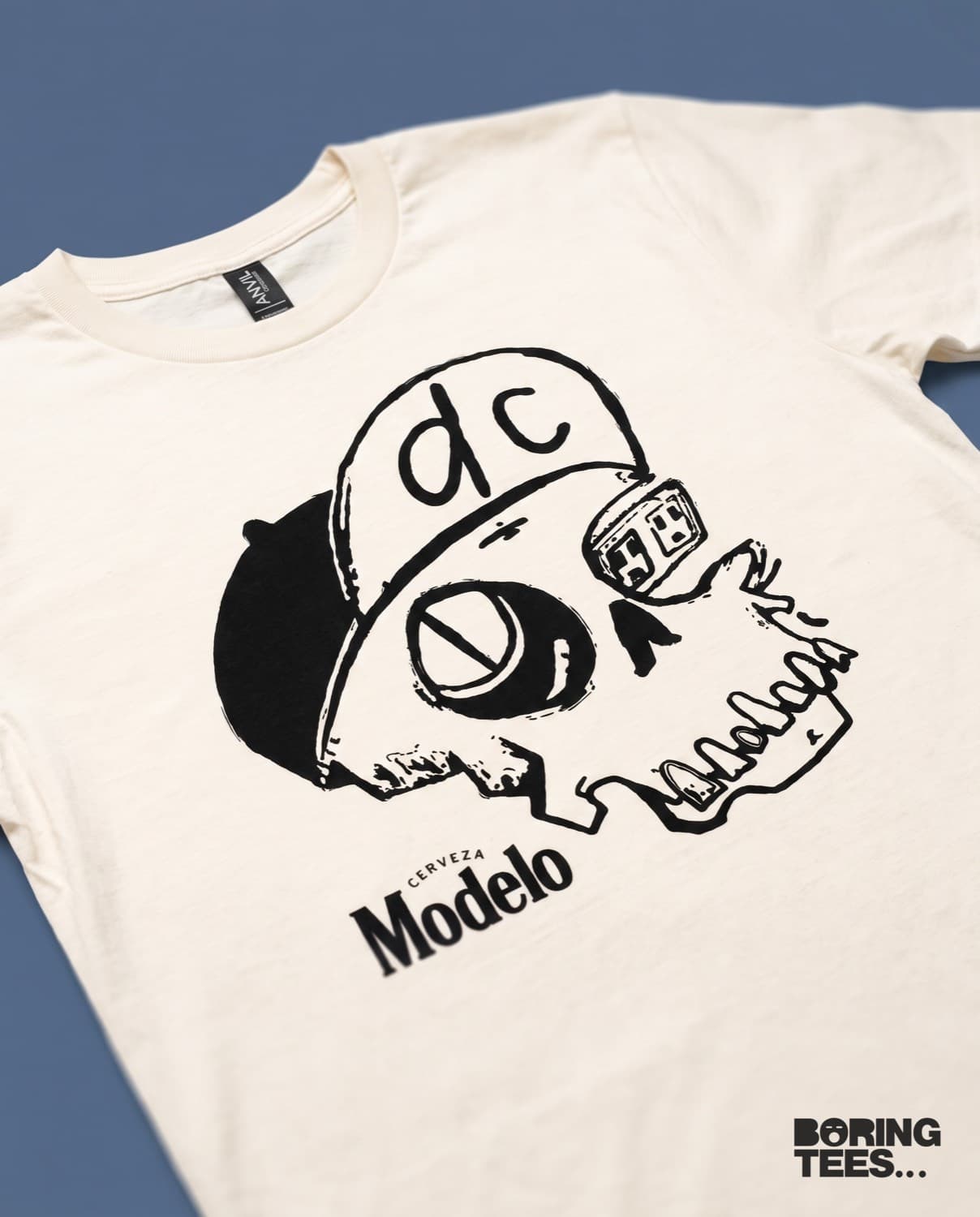 Cream tee with a black brush-drawn skull wearing a dc fitted cap above the Cerveza Modelo logo